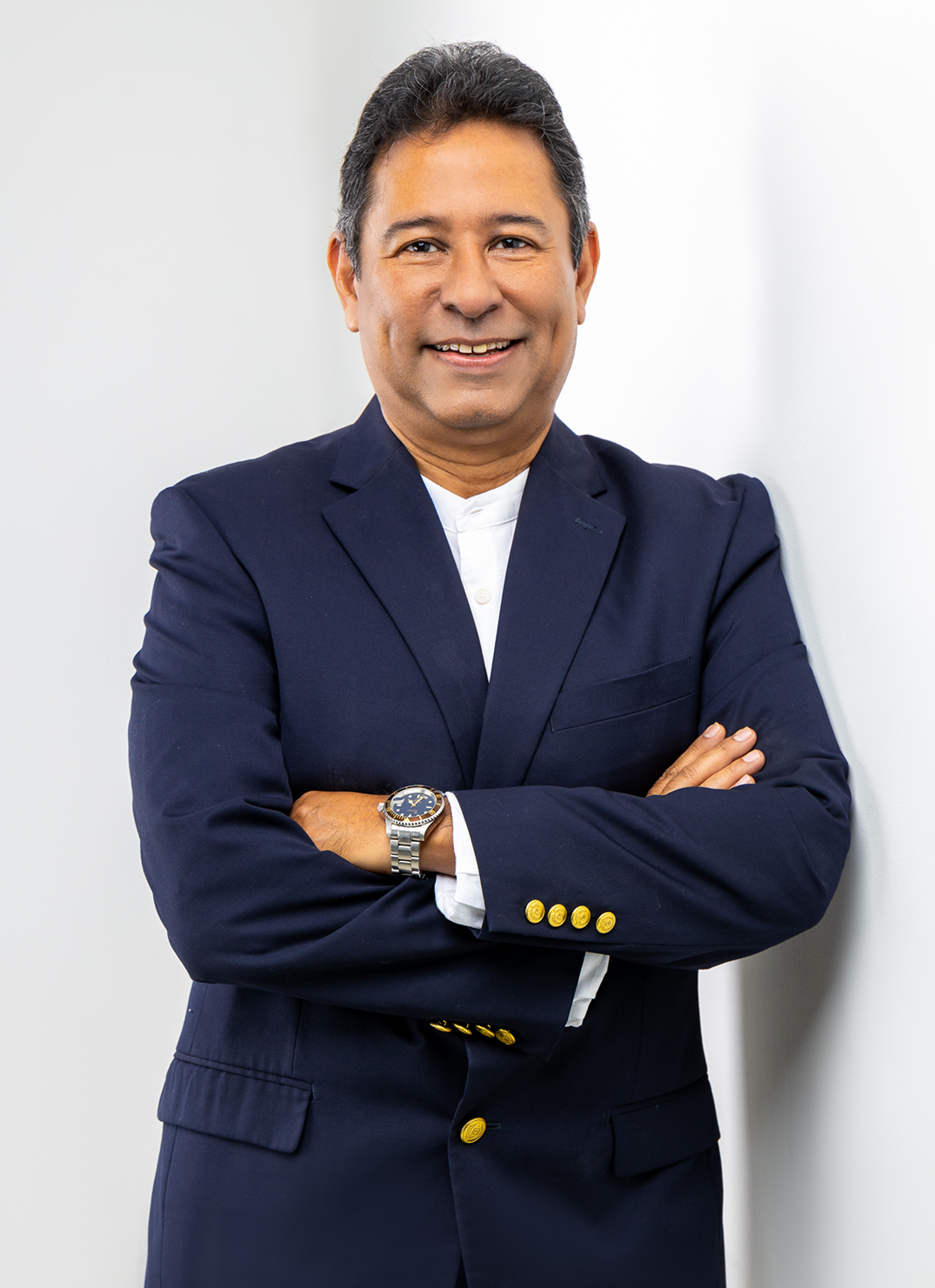 Retrato de medio cuerpo del ejecutivo Orlando Fujitsubo sonriendo con confianza y la mano en la barbilla en actitud analítica. Viste un elegante saco azul marino y un reloj clásico, destacando sobre un fondo negro sólido. La imagen transmite profesionalismo, experiencia corporativa y liderazgo empresarial.